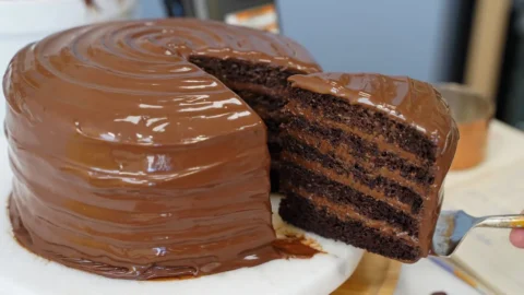 Fatia triangular de bolo de chocolate Matilda com cobertura brilhante escorrendo em um prato branco.